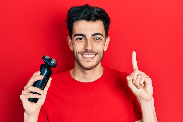 Young hispanic man holding electric razor machine smiling with an idea or question pointing finger with happy face number one