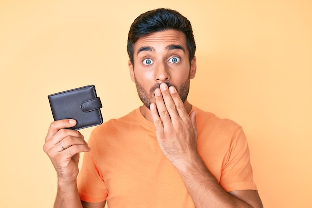 Young hispanic man holding leather wallet covering mouth with hand shocked and afraid for mistake surprised expression