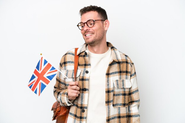 Young hispanic man holding an United Kingdom flag isolated on white background looking to the side and smiling