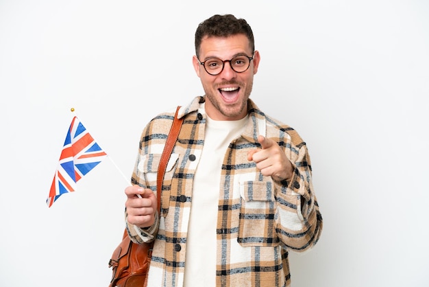 Young hispanic man holding an United Kingdom flag isolated on white background surprised and pointing front