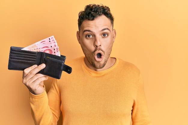 Young hispanic man holding wallet with yuan chinese banknotes scared and amazed with open mouth for surprise, disbelief face