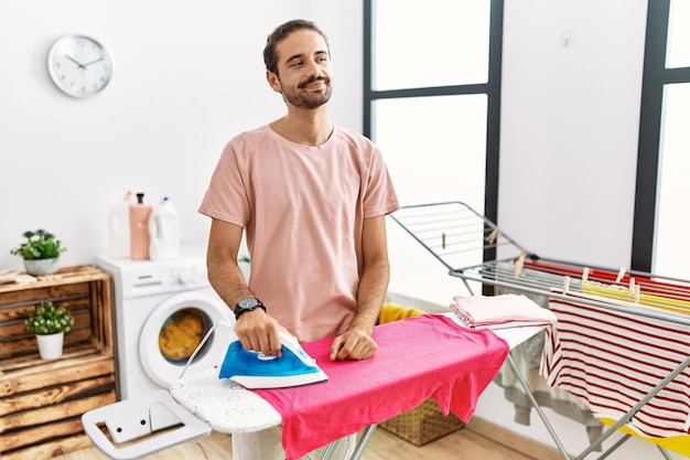 Young hispanic man ironing clothes at home looking away to side with smile on face, natural expression. laughing confident.