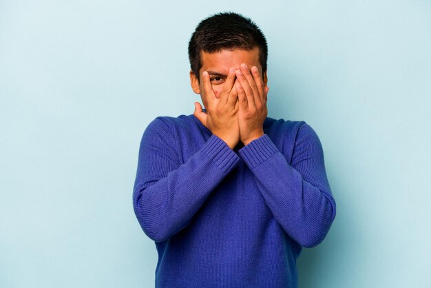 Young hispanic man isolated on blue background blink at the camera through fingers embarrassed covering face