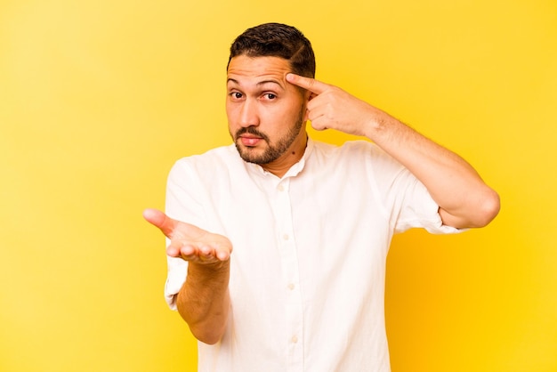 Young hispanic man isolated on yellow background holding and showing a product on hand