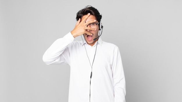 Young hispanic man looking shocked, scared or terrified, covering face with hand. telemarketer concept