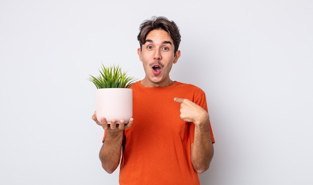 young hispanic man looking shocked and surprised with mouth wide open, pointing to self. decorative plant concept