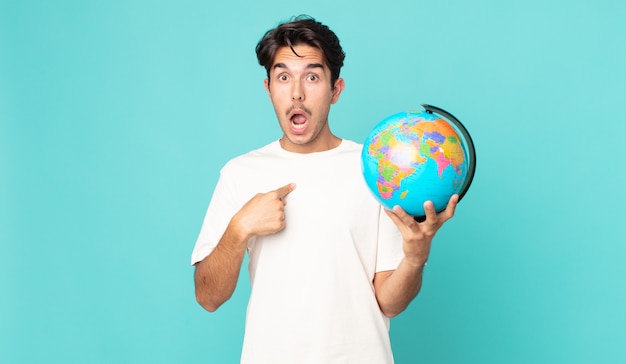 Young hispanic man looking shocked and surprised with mouth wide open, pointing to self and holding a world globe map