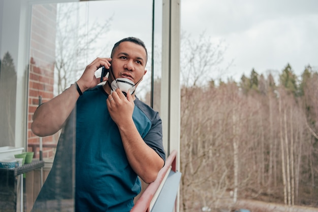 Young hispanic man in mask looking out from balcony and talks with friends on phone