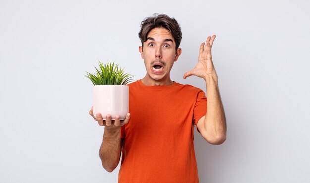 Young hispanic man screaming with hands up in the air. decorative plant concept