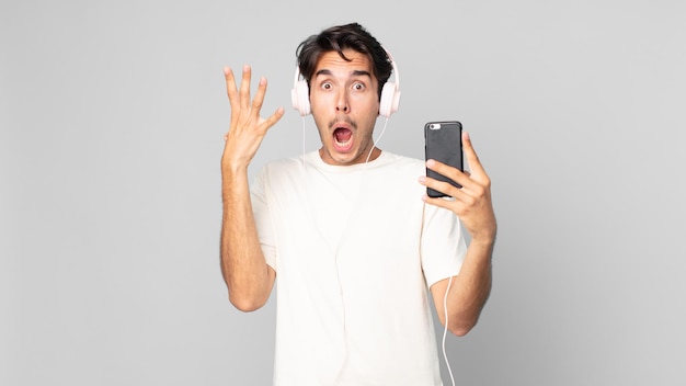 Young hispanic man screaming with hands up in the air with headphones and smartphone