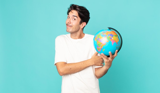 Young hispanic man shrugging, feeling confused and uncertain and holding a world globe map