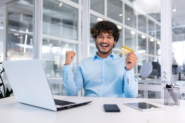 Young hispanic man sitting in the office at the desk holding a credit card looking at the camera and