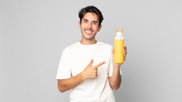 Young hispanic man smiling cheerfully, feeling happy and pointing to the side with a coffee thermos