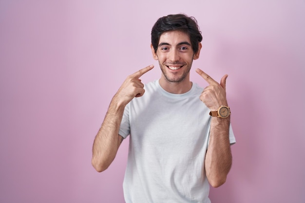 Young hispanic man standing over pink background smiling cheerful showing and pointing with fingers teeth and mouth. dental health concept.