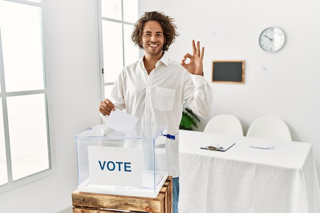 Young hispanic man voting putting envelop in ballot box smiling positive doing ok sign with hand and fingers successful expression