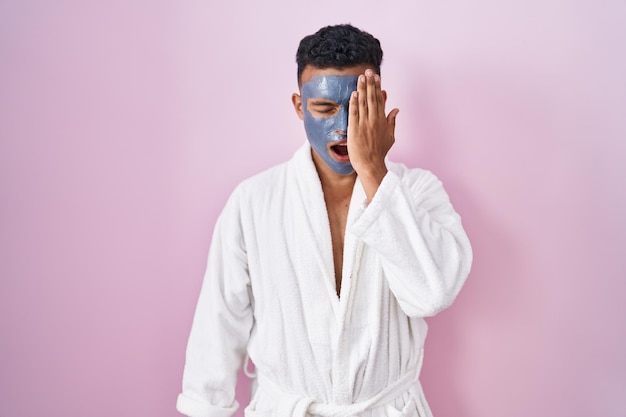 Young hispanic man wearing beauty face mask and bath robe yawning tired covering half face, eye and mouth with hand. face hurts in pain.