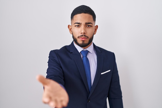 Young hispanic man wearing business suit and tie smiling cheerful offering palm hand giving assistance and acceptance.