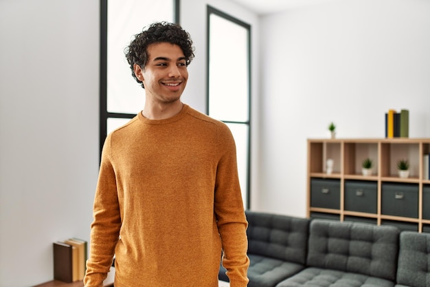 Young hispanic man wearing casual clothes standing at home looking away to side with smile on face, natural expression. laughing confident.