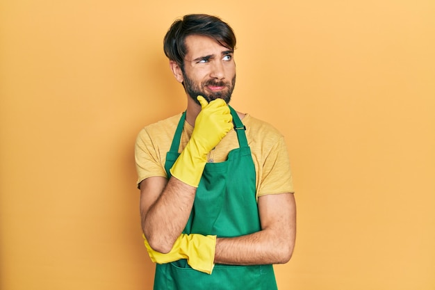 Young hispanic man wearing cleaner apron and gloves thinking concentrated about doubt with finger on chin and looking up wondering