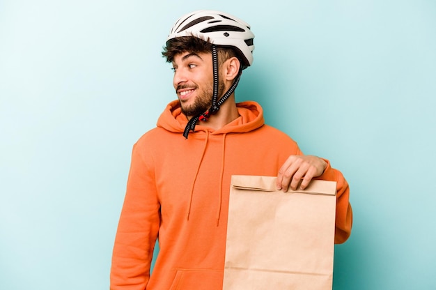 Young hispanic man wearing a helmet bike holding a take away food isolated on blue background looks aside smiling cheerful and pleasant