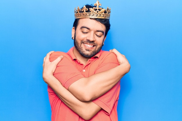 Young hispanic man wearing king crown hugging oneself happy and positive, smiling confident. self love and self care