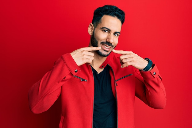 Young hispanic man wearing red leather jacket smiling cheerful showing and pointing with fingers teeth and mouth. dental health concept.