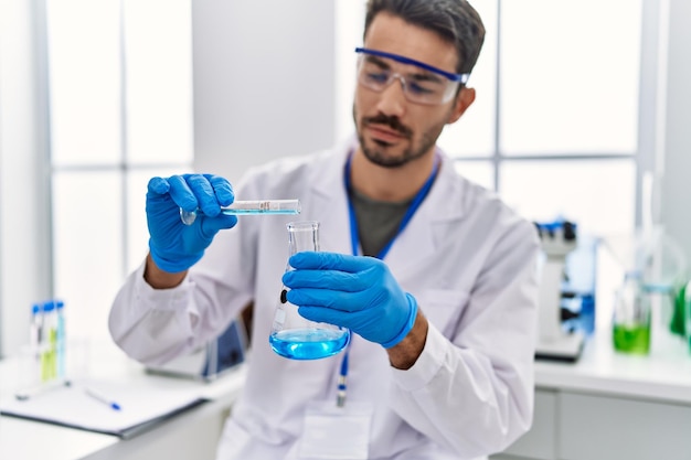 Young hispanic man wearing scientist uniform pouring liquid on test tube at laboratory