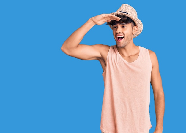 Young hispanic man wearing summer hat very happy and smiling looking far away with hand over head searching concept