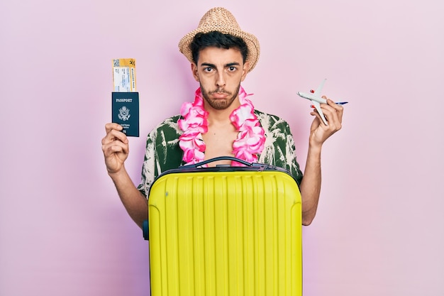 Young hispanic man wearing summer style and hawaiian lei holding passport and plane toy depressed and worry for distress crying angry and afraid sad expression