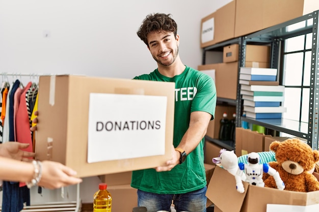 Young hispanic man wearing volunteer uniform giving donations box at charity center