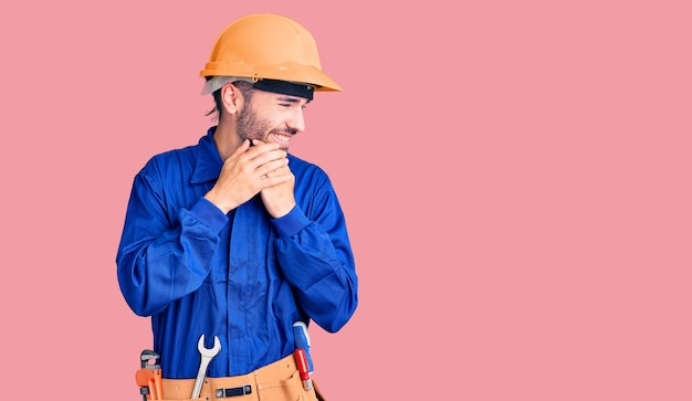 Young hispanic man wearing worker uniform laughing nervous and excited with hands on chin looking to the side