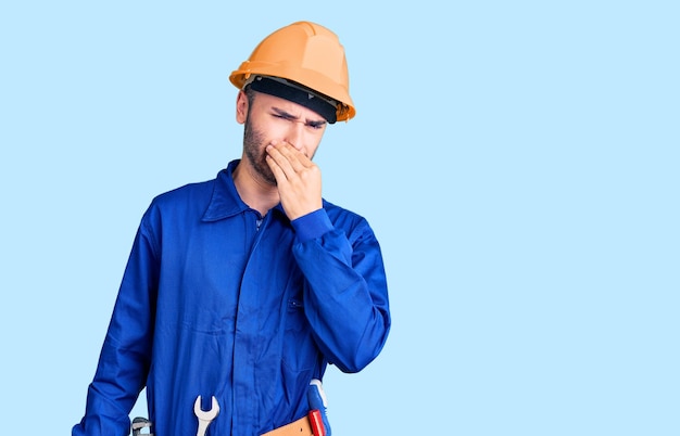 Young hispanic man wearing worker uniform smelling something stinky and disgusting intolerable smell holding breath with fingers on nose bad smell