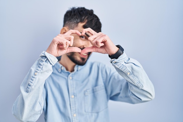 Young hispanic man with beard standing over blue background doing heart shape with hand and fingers smiling looking through sign