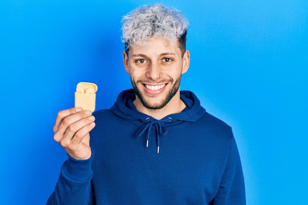 Young hispanic man with modern dyed hair holding wireless earphones looking positive and happy standing and smiling with a confident smile showing teeth