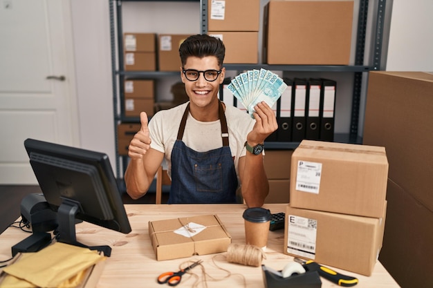 Young hispanic man working at small business ecommerce holding brazilian reals smiling happy and positive thumb up doing excellent and approval sign