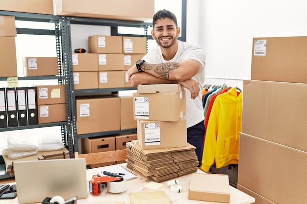 Young hispanic man working at small business ecommerce looking positive and happy standing and smiling with a confident smile showing teeth