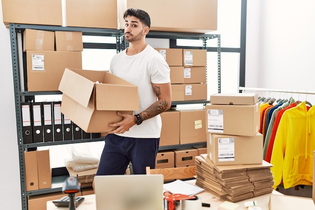 Young hispanic man working at small business ecommerce smiling looking to the side and staring away thinking.