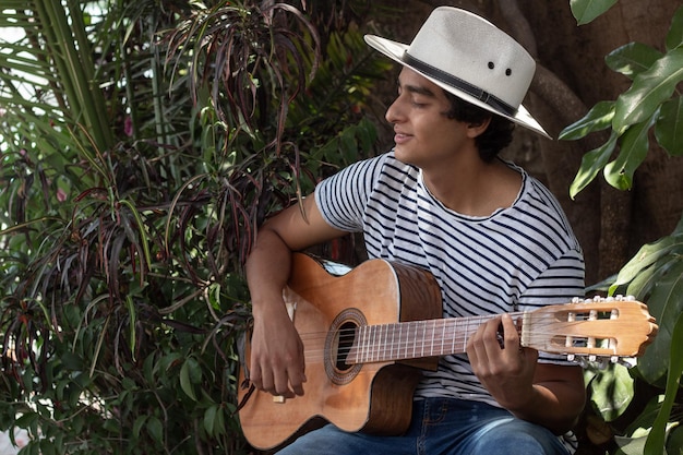 Young hispanic musician enjoys playing the acoustic guitar outdoors among the plants in the garden he enjoys his hobby very much and the expression on his face confirms it