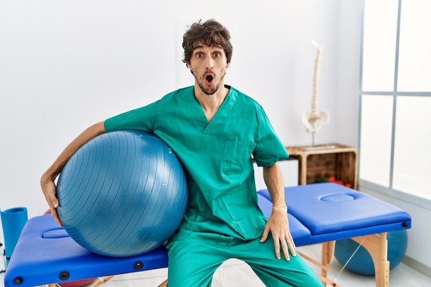 Young hispanic physiotherapist man holding pilates ball at pain recovery clinic scared and amazed with open mouth for surprise, disbelief face