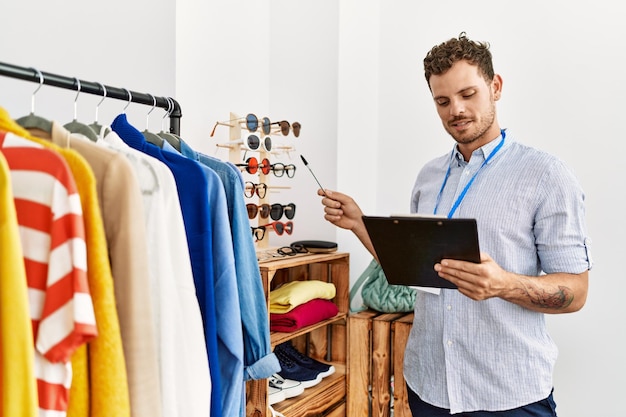 Young hispanic shopkeeper man smiling happy holding clipboard working at clothing store