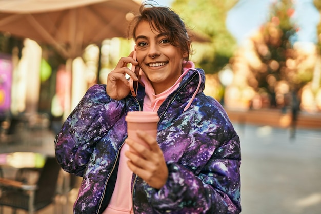 Young hispanic sporty woman talking on the smartphone drinking coffee at the city