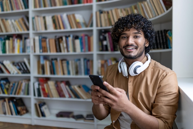 Young hispanic student inside public library studying smiling and looking at camera man holding