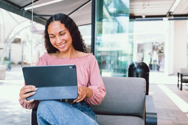 Young hispanic student woman smiling and using a tablet sitting outside a cafe in her free time