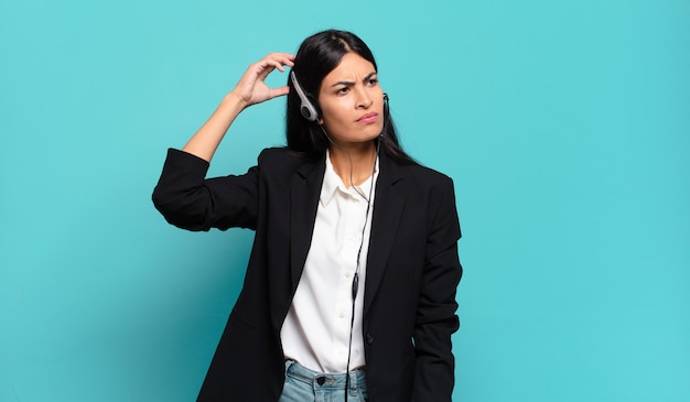 Young hispanic telemarketer woman feeling puzzled and confused, scratching head and looking to the side