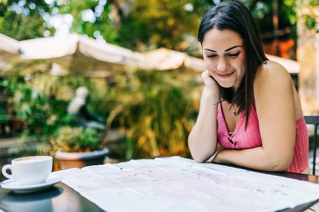 Young hispanic tourist looking a map while sitting on a cafe. Trendy woman using a smart phone while traveling alone and looking for new local places - Summer holidays and travel Europe concept