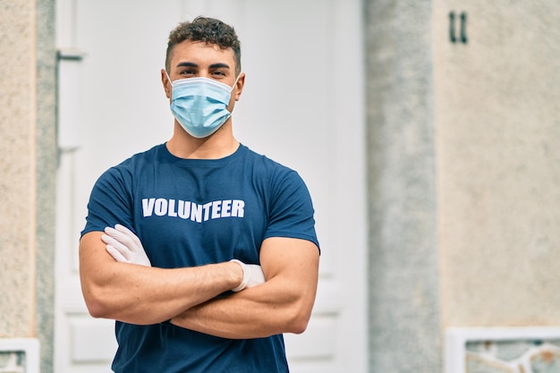 Young hispanic volunteer man with arms crossed wearing medical mask at the city.