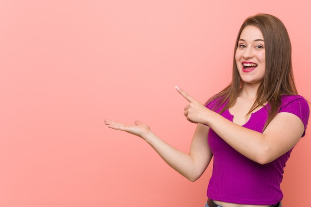 Young hispanic woman against a pink wall excited holding a copy space on palm.