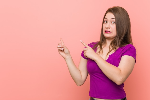 Young hispanic woman against a pink wall shocked pointing with index fingers to a copy space.