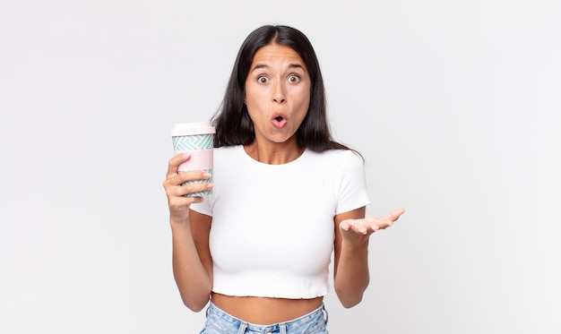 Young hispanic woman amazed, shocked and astonished with an unbelievable surprise and holding a take away coffee container