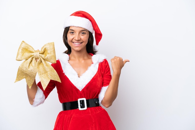 Young hispanic woman dressed as mama noel holding a Christmas bow isolated on white background pointing to the side to present a product